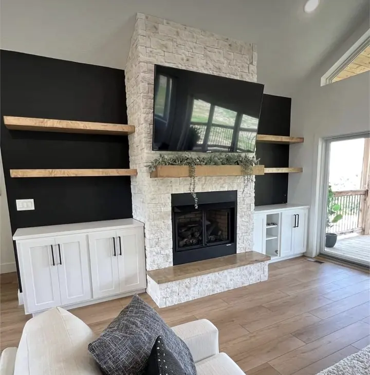 Stacked stone fireplace wall flanked by dark walls, white built-in cabinets and floating wood shelves
